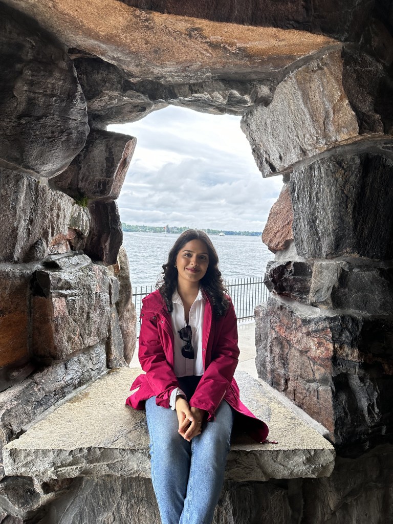 A young woman with dark, wavy hair sits on a stone bench framed by a rustic rock archway. She is wearing a burgundy jacket over a white blouse and blue jeans, with sunglasses hanging from her shirt. She smiles gently at the camera with her hands clasped on her lap. Behind her, a body of water stretches toward the horizon, with a cloudy sky and distant trees visible in the background. A black metal fence lines the waterfront.