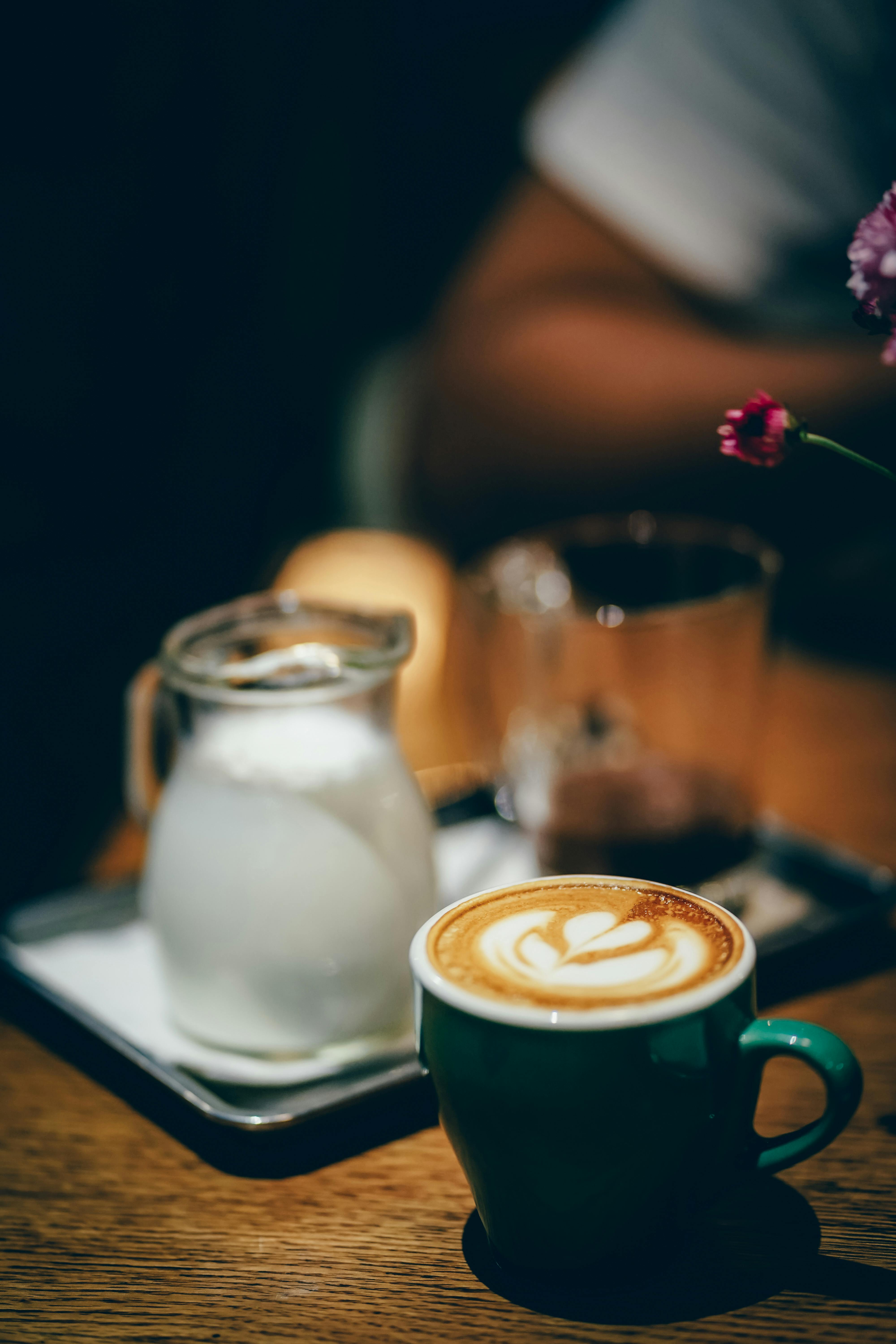 A cozy café scene with a cup of cappuccino featuring a latte art design on a wooden table. A small glass pitcher of milk and a blurred coffee setup are in the background.