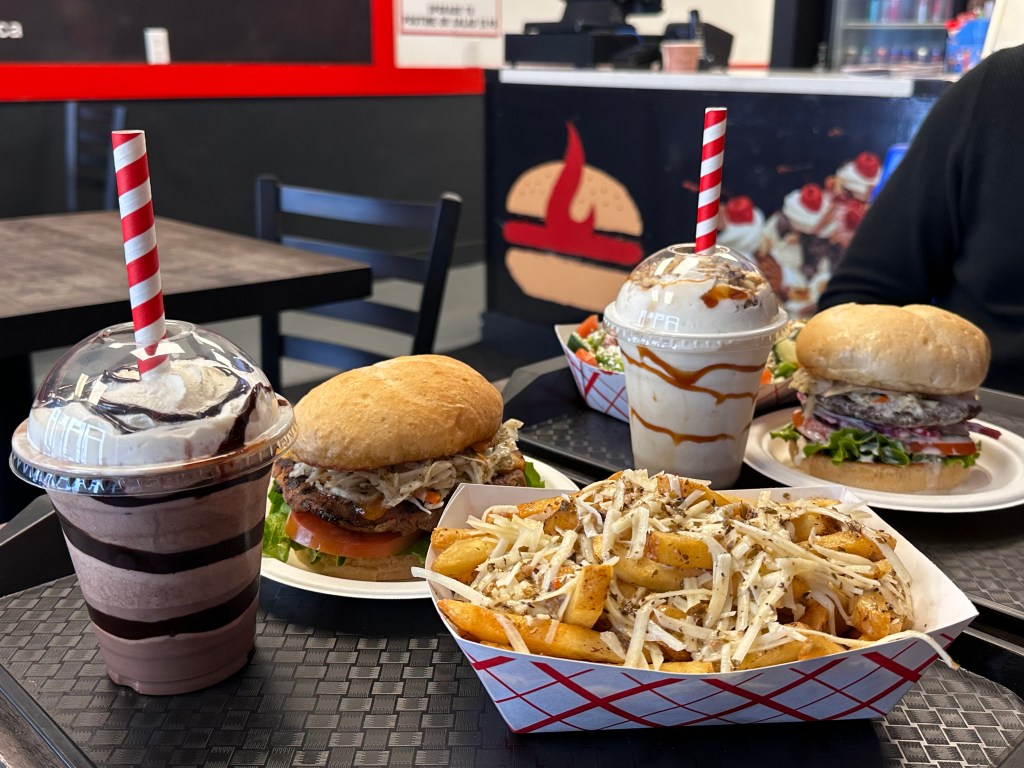 The image showcases a delicious meal from a restaurant, featuring a chocolate milkshake with whipped cream and a red-and-white striped straw on the left. To the right, there are two large burgers with fresh toppings, including lettuce, tomato, and shredded coleslaw. In the center, there is a generous serving of crispy fries topped with shredded cheese. The food is served on a black tray with a grid design, and in the background, the restaurant's interior features a black and red theme with a burger logo on the wall.