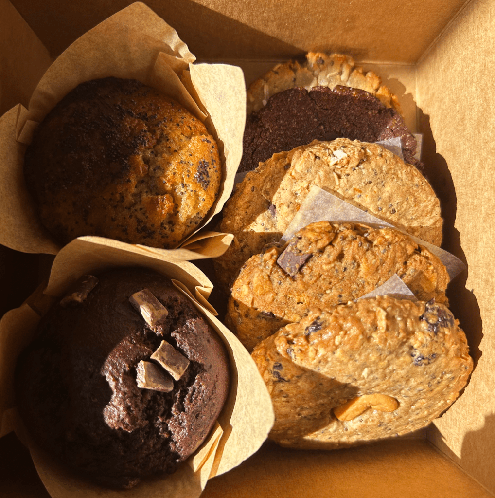 A box filled with assorted baked goods, including muffins and cookies, wrapped in parchment paper. The items include a chocolate muffin with chunks of chocolate, a poppy seed muffin, and several cookies with various textures and ingredients, like oats and chocolate chunks.