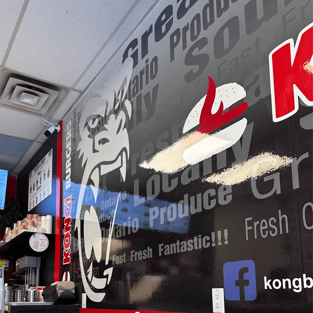 The image shows a wall mural inside a restaurant, featuring a large graphic of a gorilla's face with sharp fangs and a prominent red flame symbol. The wall is decorated with words like "Fresh," "Locally," "Ontario," "Produce," and "Fantastic," creating a bold and dynamic look. The restaurant's branding, "KONG BURGERS," is displayed in red along the side, with a Facebook link at the bottom. The atmosphere appears casual and modern, with cups stacked on the counter in the foreground.